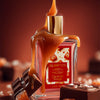 Glampton Caramel Rouge Shimmer Fragrance Mist bottle covered in dripping caramel, surrounded by chocolate pieces on a warm caramel-toned background.