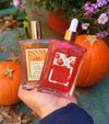 Hand holding Glampton Shimmer Fragrance Mist bottles, Vanilla Sun Musk and Shimmering Body Oil Caramel Rouge, with pumpkins, autumn leaves, and flowers in the background.