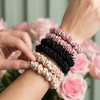 Six hair scrunchies (cream, black, pink) being worn on a wrist with flowers in the background.